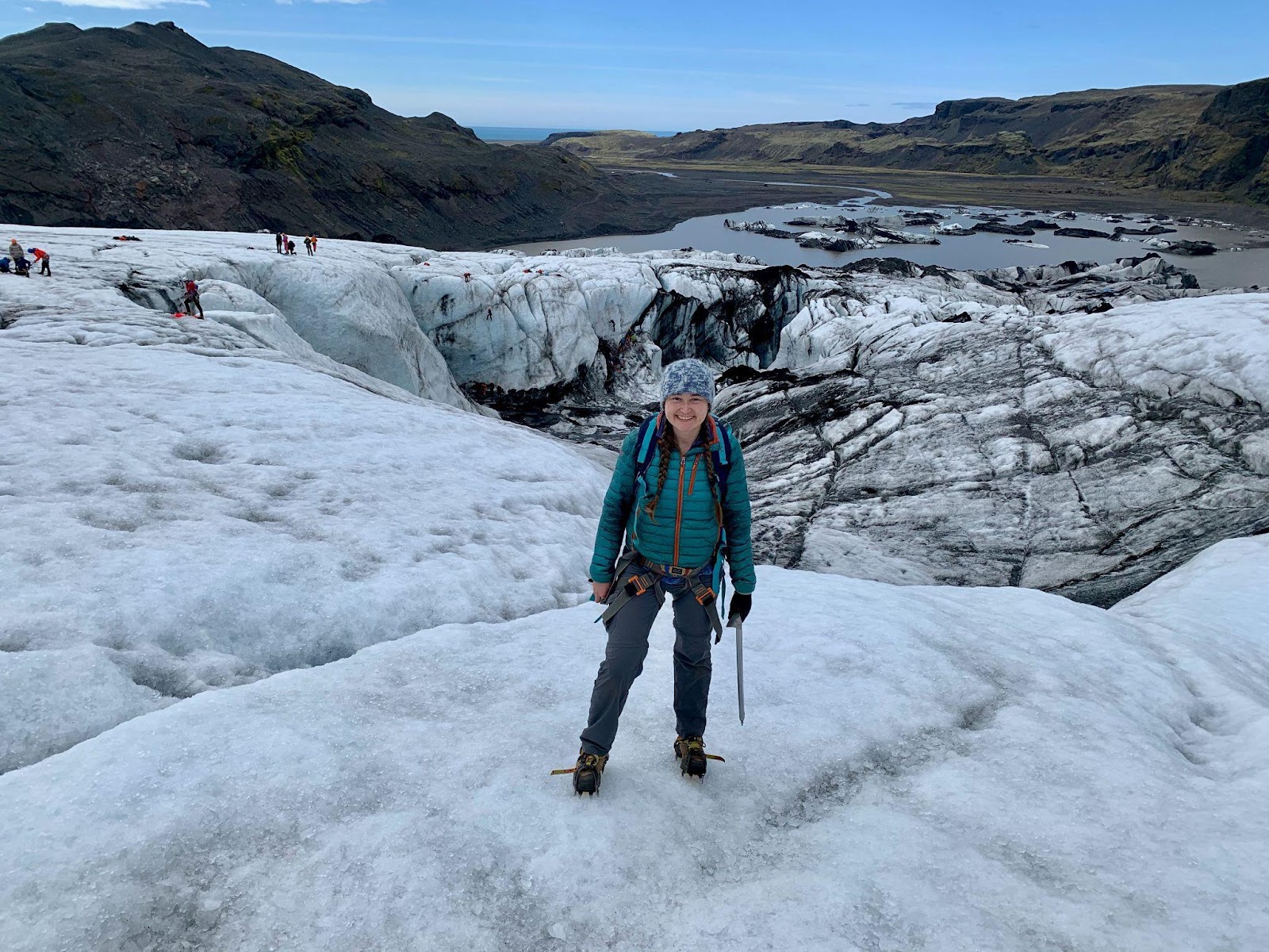 A woman stands on a glacier. Behind her, the ice gives way to a stream of water that weaves between two sets of hills into the distance.